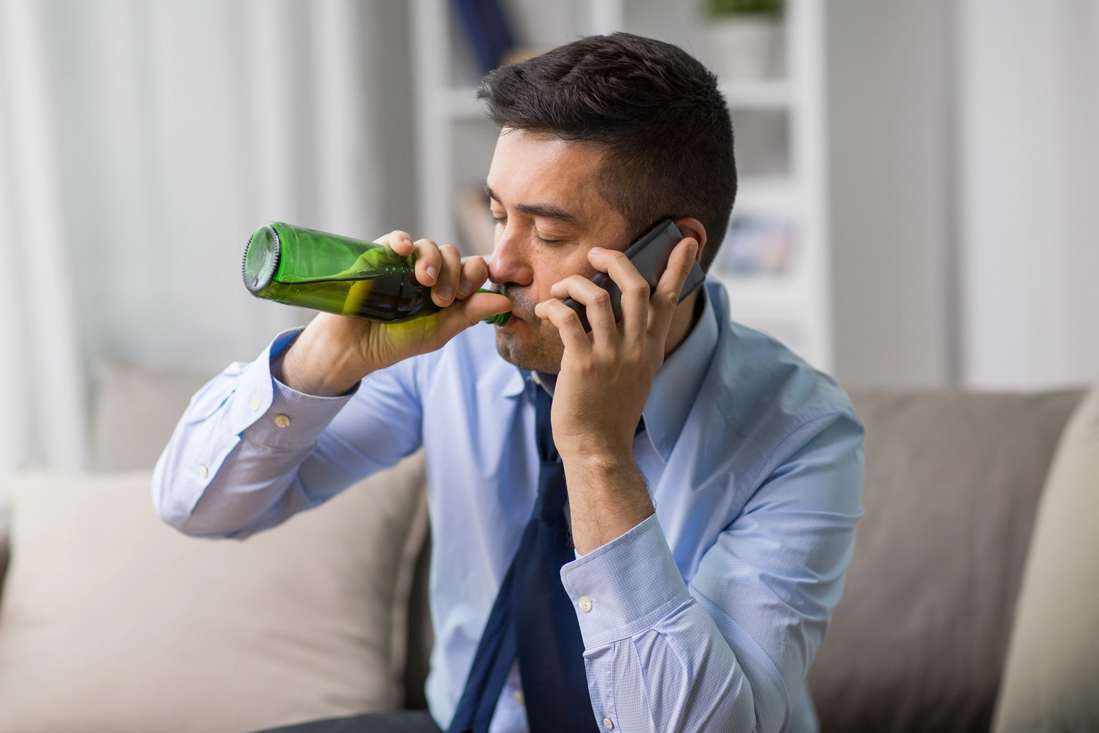 Alcoholism in Men: A man in business attire sitting on a couch drinking a beer while on the phone.