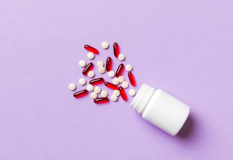 A white pill bottle on its side with red and white pills spilling across a pale purpose background.