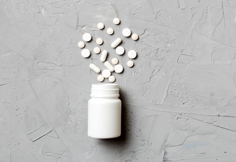 A white pill bottle with white pills of various shapes spread around on a gray surface.